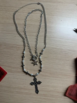 Dubbelhalsband med kors och pärlor - Snyggt dubbelhalsband i silverfärgad metall med ett stort korshänge och vita pärlor. Ena kedjan har ett stjärnhänge och små detaljer, medan den andra har pärlor och kors. Perfekt för dig som gillar statement-smycken och lager på lager.