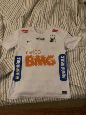 Vit Santos Neymar Jr matchtröja - Vit fotbollströja från Nike för Santos FC med Neymar Jr och nummer 11 på ryggen. Tröjan har flera sponsorloggor i orange, blått och svart samt klubbmärke på bröstet. Korta ärmar och rund hals, tillverkad i lätt och ventilerande material.    Köpt på plick