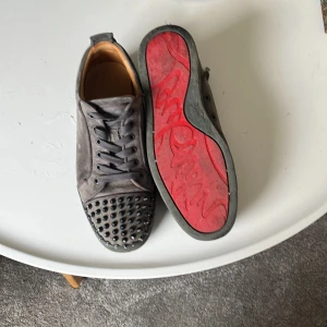Christian Louboutin - Snygga grå sneakers i mocka från Louboutin med ikonisk röd sula och svarta nitar på tån. Skorna har snörning och en rund tå, vilket ger en edgy och lyxig vibe. Perfekta för dig som vill sticka ut med exklusiva detaljer och streetstyle-känsla. Piggarna är lite slitna på ena skon och det är däremot fyra piggar borta på vänster skon vilket går att köpa på Louboutin hemsidan för runt 200 kr