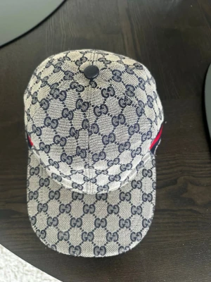 Gucci keps med GG-mönster - Snygg keps från Gucci i beige och marinblått med klassiskt GG-monogram över hela kepsen. Justerbar rem baktill i läder och diskret röd-blå rand på sidan. Tillverkad i canvasmaterial med exklusiv känsla.