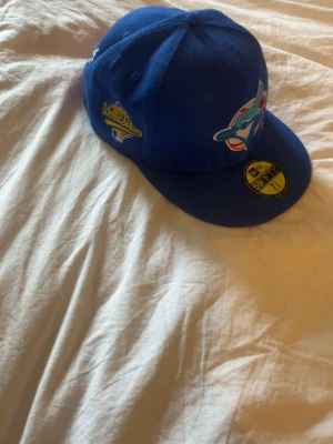 Blå Toronto Blue Jays keps - Säljer en blå New Era 59FIFTY keps med Toronto Blue Jays-logga framtill och World Series-märke på sidan. Klassisk flat brim och broderade detaljer. Perfekt för dig som gillar sportig stil och vill sticka ut med en ikonisk lagkeps.