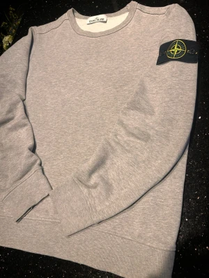 Grå sweatshirt från Stone Island - S  - Snygg grå sweatshirt från Stone Island med klassisk rund hals och ribbade muddar. Tröjan har den ikoniska svarta patchen med gult och grönt Stone Island-märke på vänster ärm. Tillverkad i mjuk bomullsblandning för skön komfort.