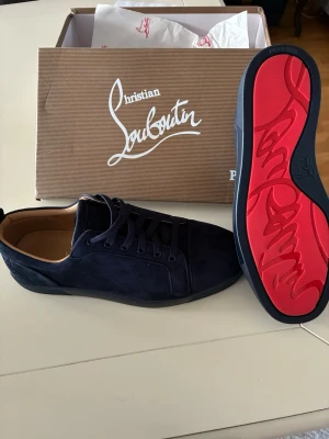 Louboutin - Snygga mörkblå sneakers från Christian Louboutin i mocka med klassisk röd sula och diskret logga på sidan. Skorna har låga skaft, svarta skosnören och en lyxig känsla med beige innersula. Perfekta för dig som vill sticka ut med exklusiv stil.