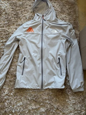 Ljusgrå vindjacka från The North Face - Snygg ljusgrå vindjacka från The North Face med huva och hel dragkedja. Jackan har två framfickor med dragkedja, elastiska muddar och logga på bröstet samt ryggen. Materialet är tunt och smidigt, perfekt för blåsiga dagar. Orange detalj på bröstet för extra stil.