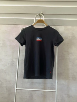 Svart Levi's t-shirt med logga - Snygg svart t-shirt från Levi's med klassisk logga i vitt, blått och rött på bröstet. T-shirten har rund halsringning och korta ärmar. Tillverkad i mjuk bomull som känns skön mot huden. Perfekt till jeans eller shorts för en avslappnad look. (Aldrig använd, men saknar prislapp)
