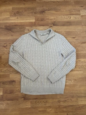 Beige kabelstickad tröja Only & Sons - Snygg beige kabelstickad tröja från Only & Sons med halv dragkedja vid kragen. Tröjan har långa ärmar, ribbade muddar och en klassisk kabelstickad design som ger en stilren look. Perfekt för lager på lager och passar till det mesta. 