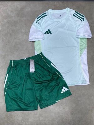 Adidas grön och vit träningsset - Snyggt träningsset från Adidas med en vit t-shirt med gröna detaljer och gröna shorts med vita inslag. Perfekt för fotboll eller annan träning. Setet har klassiska Adidas-loggor och är gjort i ett lätt och ventilerande material.