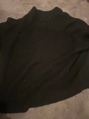 Svart stickad polotröja - En klassisk svart stickad polotröja med hög krage och lång ärm. Perfekt för lager-på-lager och enkel att matcha med olika outfits. Tröjan har en rak passform och ribbstickade detaljer vid hals, ärmslut och nederkant.