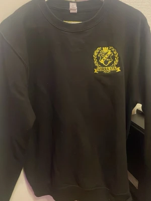 Svart AIK crewneck med Norra Stå-tryck - Svart crewneck-tröja med AIK:s emblem och texten 'Norra Stå' i gult på bröstet. Baksidan har ett schackrutigt gult tryck i nacken. Klassisk passform och rund halsringning, perfekt för dig som vill visa ditt AIK-stöd.