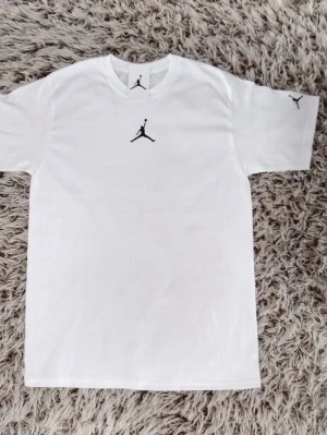 Vit Jordan t-shirt med logga - Säljer en clean vit t-shirt från Jordan med den klassiska svarta Jumpman-loggan på bröstet och ärmen. T-shirten har normal passform, rund halsringning och är tillverkad i mjuk bomull. Perfekt för en sportig och avslappnad stil.