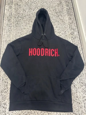 Svart HOODRICH hoodie med röd logga - Svart hoodie från HOODRICH med stor röd logga på bröstet. Klassisk modell med huva och dragsnören samt en stor magficka. Tillverkad i mjukt material som passar perfekt för en avslappnad streetwear-look.