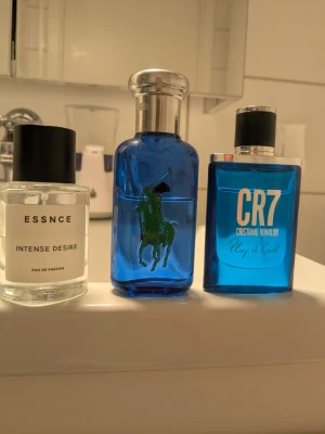 Tre parfymer: Essnce, Ralph Lauren, CR7 - Säljer tre parfymer: Essnce Intense Desire i genomskinlig flaska med svart lock, Ralph Lauren Polo Blue 1 i blå glasflaska med silverfärgat lock och gul siffra, samt CR7 Play It Cool av Cristiano Ronaldo i blå flaska med svart och silverfärgat lock.