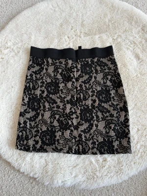 Svart spetsmönstrad kjol H&M Divided (XS) - Kort kjol från H&M Divided med svart spetsmönster över beige botten. Kjolen har bred resår i midjan och ett elegant blommigt mönster som ger en cool och trendig look. Perfekt att styla med en enkel topp och boots.