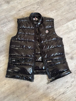 Svart dunväst från Moncler - Snygg svart dunväst från Moncler med glansig finish och klassisk logotyp på bröstet. Västen har två dragkedjeförsedda fickor framtill och quiltad design. Perfekt för lager-på-lager och har coolt innerfoder med serietidningsmotiv.