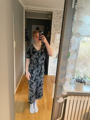 Svart jumpsuit med palmtryck - Cool svart jumpsuit med vitt palmbladsmönster. För mig är den för kort men borde passa någon kortare än jag perfekt. V-ringad framtill, korta ärmar och knytband i midjan för en snygg siluett. Nyckelhålsöppning i ryggen. Materialet känns mjukt och lätt, perfekt för sommaren. 