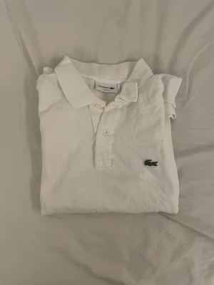 Vit pikétopp från Lacoste - Vit pikétopp från Lacoste med klassisk krage, korta ärmar och knappar framtill. Toppen är i bomull och har den ikoniska gröna krokodilloggan broderad på bröstet. Enkel och stilren design som ger en sportig känsla.