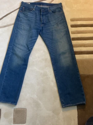 Levi's 501 blå jeans W34 L30 - Klassiska blå Levi's 501 jeans med rak passform och knappgylf. Jeansen är i 100% bomull och har den ikoniska läderpatchen bak. Perfekta för en avslappnad och tidlös stil.
