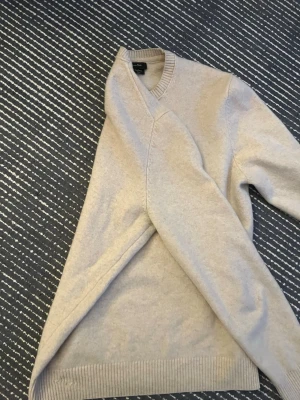 Beige stickad tröja  - Beige stickad tröja, med rund halsringning och ribbade muddar vid ärmslut och nederkant. Tröjan har en klassisk och stilren design, perfekt för lager-på-lager och enkel att matcha med olika outfits. Materialet känns mjukt och bekvämt. Den har små hål och små fläckar men inget som syns om man inte aktivt letar efter det.