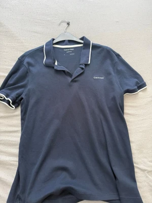 Mörkblå pikétröja från Calvin Klein - Snygg mörkblå pikétröja från Calvin Klein med vit kant på krage och ärmslut. Klassisk passform med korta ärmar och liten logga broderad på bröstet. Perfekt för en stilren look.
