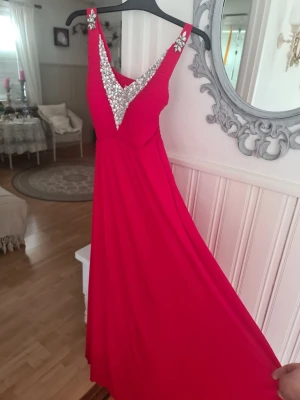Röd långklänning med paljetter - Snygg röd långklänning med smala axelband och v-ringning dekorerad med silvriga paljetter. Klänningen har ett elegant fall och markerad midja, perfekt för bal eller fest. Materialet känns mjukt och följsamt.Det saknas en sten på klänningen som kan ses på bilden.