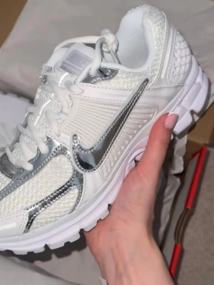 Nike Vomero Zoom 5 Chrome - Nike sneakers i vitt med silvriga detaljer och meshpaneler. Skorna har en chunky sula och klassisk snörning. Loggan i metallic silver. Perfekta för dig som gillar sportig och trendig stil.