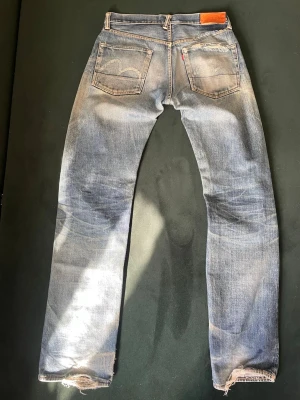 SKIT FETA EVISU - Säljer ett par klassiska blå evisu jeans med raka ben, slitningar och hål på vänster knä samt fransiga benslut. Jeansen har fem fickor, läderpatch bak och den ikoniska röda Levi's-lappen. Perfekt för en avslappnad och trendig look.