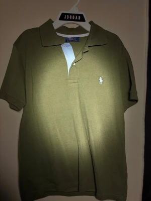 Grön piké från Polo Ralph Lauren - Snygg grön pikétröja från Polo Ralph Lauren med klassisk krage och korta ärmar. Tröjan har den ikoniska broderade loggan i blå på bröstet och en kontrastdetalj i ljusblått vid knäppningen. Perfekt för en avslappnad men stilren look.