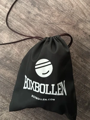 Boxbollen träningsboll med pannband - Säljer en Boxbollen träningsboll med svart elastiskt pannband och röd boll. Perfekt för reflex- och boxningsträning. Kommer i en svart förvaringspåse med vit logga. Enkel att ta med och använda både inne och ute. Om repet går sönder kan man lägga ett annat rep så att man kan fortsätta använda den. Väldigt bra för att träna sina slag på en boll