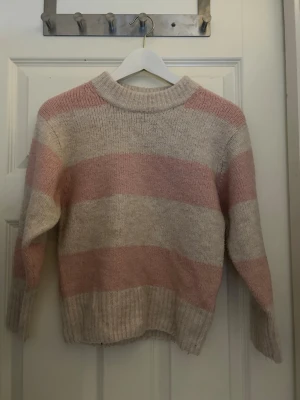 Stickad tröja med breda ränder - Mysig stickad tröja med breda horisontella ränder i ljusrosa och beige. Tröjan har en hög, ribbad halsringning och långa ärmar med ribbade muddar. 