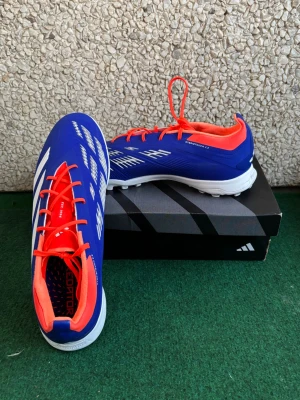 Elite Fotbollsskor(påse ingår) - Säljer ett par Adidas Predator fotbollsskor i blått med vita detaljer och röda snören. Skorna har en låg profil, rund tå och platt sula med dobbar för bra grepp på konstgräs. Ovandelen är i syntetmaterial och insidan har en röd kontrast. Perfekt för dig som vill sticka ut på planen.  Skickar även med en gratis påse som du ser på bild 5. Den är inte så mycket använt, men bara att konstgräs färgen stanna kvar. Den är för Konstgräs,Turf, utomhus skor. I