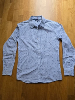 Blå randig skjorta från Tommy Hilfiger - Säljer en snygg blå skjorta från Tommy Hilfiger med tunna vita ränder och små röda och vita logomönster. Skjortan har klassisk krage, knappar framtill och långa ärmar. Perfekt för dig som gillar stilrena detaljer och ikoniskt märke. Skjortan är ny utan prislapp str s elle 170. Ungdon