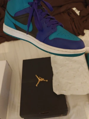 Jordan 1 mid black grape  - Säljer ett par Jordan 1 High sneakers i en cool mix av blått, lila och svart med vita sulor. Skorna har klassisk hög siluett, lädermaterial och ikonisk swoosh på sidan. Perfekta för dig som vill sticka ut med färg och stil.3900 NY PRIS 