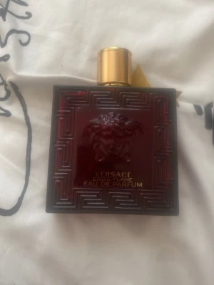 Versace Eros Flame röd parfym - Versace Eros Flame parfym i en snygg, fyrkantig röd glasflaska med guldfärgat lock och Medusa-detalj. Flaskan har ett elegant mönster och tydlig logga framtill. Perfekt för dig som gillar exklusiva och stilrena dofter.