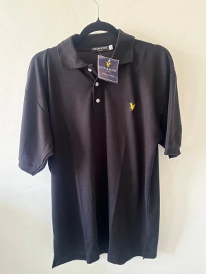 Svart pikétröja från Lyle & Scott. Passar även L - Klassisk svart pikétröja från Lyle & Scott med korta ärmar och gul logga broderad på bröstet. Tröjan har krage och knappar framtill. Perfekt för en stilren och avslappnad look.