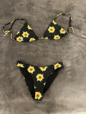 Svart bikini med solrosor - Snygg svart bikini med gula solrosor och trekantsmodell på överdelen. Triangel kuporna är justerbara år sidorna, både inåt och utåt. Och även axelbanden är justerbara, vilket gör det lätt att anpassa toppens passform. Perfekt för dagar på stranden eller poolhäng. Hör av er vid fler frågor 