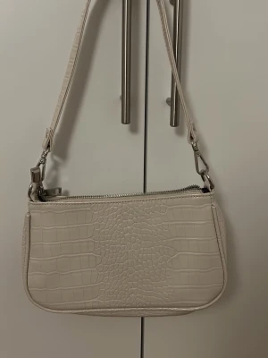 Beige axelremsväska i krokopräglat skinn - Snygg beige handväska med axelrem och krokodilpräglat mönster. Väskan har silverfärgade metalldetaljer och dragkedja upptill. Perfekt storlek för det viktigaste och ger en trendig touch till din outfit.