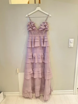 Ljuslila maxiklänning/balklänning True Decadence - Drömmig långklänning i ljuslila från True Decadence. Klänningen har tunna axelband, volangdetaljer i flera lager och spets vid midjan. Tillverkad i ett lätt, flowy material som ger en romantisk vibe. Perfekt för speciella tillfällen.