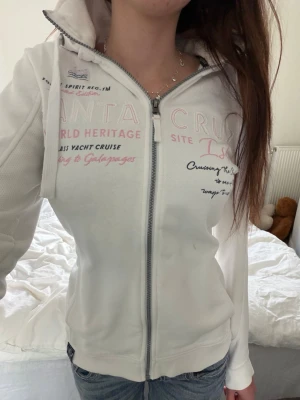 Zip hoodie från Geographical Norway - Vit hoodie med dragkedja från Geographical Norway. Hoodien har broderade och tryckta detaljer i rosa och svart på bröstet och ärmen, samt texten 'Santa Cruz' och 'World Heritage'. 