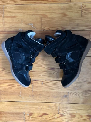 Svarta sneakers med kardborre och kilklack - Säljer ett par svarta sneakers med kilklack och coola kardborreband. Skorna har en chunky brun sula och är gjorda i mocka och skinn. De har högt skaft och detaljer i svart och grått som ger en sportig vibe.