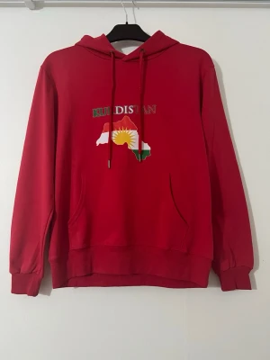 Röd hoodie med Kurdistan-tryck - Röd hoodie med huva, samt tryck av Kurdistan-karta och flagga på både fram- och baksidan. Stora fickor framtill och ribbade muddar vid ärmslut och nederkant. Perfekt för dig som vill visa ditt stöd. Priset kan diskuteras 