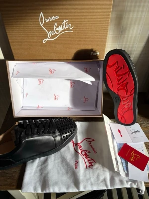 L - Säljer ett par svarta sneakers från L i läder med ikoniska röda sulor och svarta nitar på tån. Skorna har klassisk låg profil och svarta skosnören. Kommer med originalkartong, dustbag och certifikat.