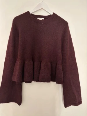 Vinröd stickadtröja - Mysig vinröd stickad tröja med volang i midjan och vida ärmar. Tröjan har rund halsringning och är tillverkad i ett mjukt, fluffigt material. Mycket bra skick använd endast 2 gånger.