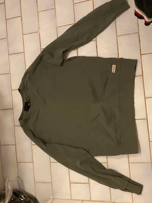 Olivgrön sweatshirt från 157 - Olivgrön sweatshirt från 157 i storlek M. Klassisk rund halsringning och ribbade muddar vid ärmslut och nederkant. Perfekt basictröja med diskret logga nertill. Skön och avslappnad passform.
