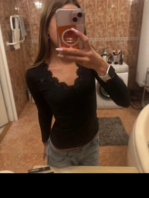 Svart långärmad topp med spets - Snygg svart långärmad topp med v-ringning och spetsdetaljer runt halsen. Figurnära passform och stretchigt material som sitter bekvämt. Perfekt att matcha med jeans eller kjol för en trendig look.