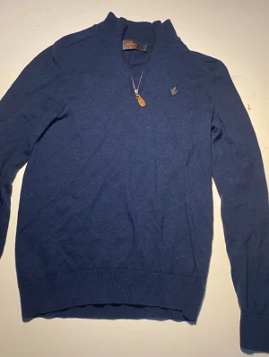 Mörkblå stickad half zip tröja från Morris,  - 100% Merino Ull, Mörkblå stickad tröja från Morris med halv dragkedja och liten broderad logga på bröstet. Tröjan har ribbade muddar vid ärmslut och nederkant samt hög krage.  Perfekt för lager på lager och snygg till jeans. Det står att det är storlek M men passar mer som storlek S. 