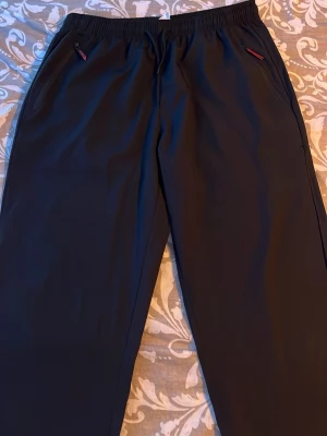 Svarta mjukisbyxor med dragkedjor - Säljer ett par svarta mjukisbyxor med elastisk midja och dragkedjor på fickorna. Byxorna har en rak passform och är tillverkade i ett mjukt och bekvämt material, perfekta för chill dagar. Snygga detaljer med röda dragkedjor på fickorna.