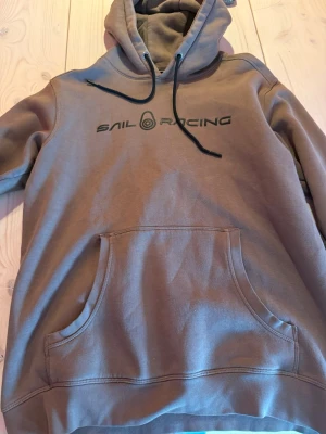 Brun hoodie från Sail Racing - Säljer en brun hoodie från Sail Racing med svart logga på bröstet. Hoodien har huva med svarta dragsnören och en stor magficka framtill. Tillverkad i mjukt bomullsmaterial, perfekt för chill dagar och streetwear-stil. Fint skick 