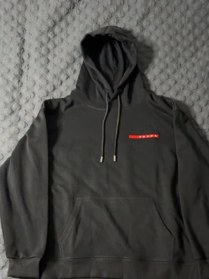 Svart hoodie från Prada  - Snygg svart hoodie från prada med klassisk röd logga på bröstet. Hoodien har dragsko i huvan och en stor magficka framtill. Tillverkad i mjukt bomullsmaterial som känns skönt mot huden. Perfekt för dig som gillar stilrena designer och exklusiva märken. STORLEK M, PASSAR M OCH S, HELT NY, PRISLAPP PÅ!!