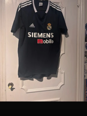Real Madrid bortatröja Adidas - Mörkblå Real Madrid bortatröja från Adidas med Siemens Mobile som sponsor. Tröjan har vita detaljer, klubbmärke på bröstet och LFP-märke på ärmen. Klassisk fotbollströja med v-ringning och tre vita ränder på axlarna.