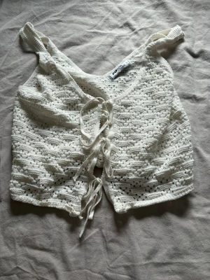 Vit virkad linnetopp med snörning - Söt vit virkad linnetopp med v-ringning och snörning framtill. Toppen är ärmlös och har ett luftigt, somrigt material som passar perfekt till varma dagar. Stilsäker detalj med knyt framtill för en trendig look.